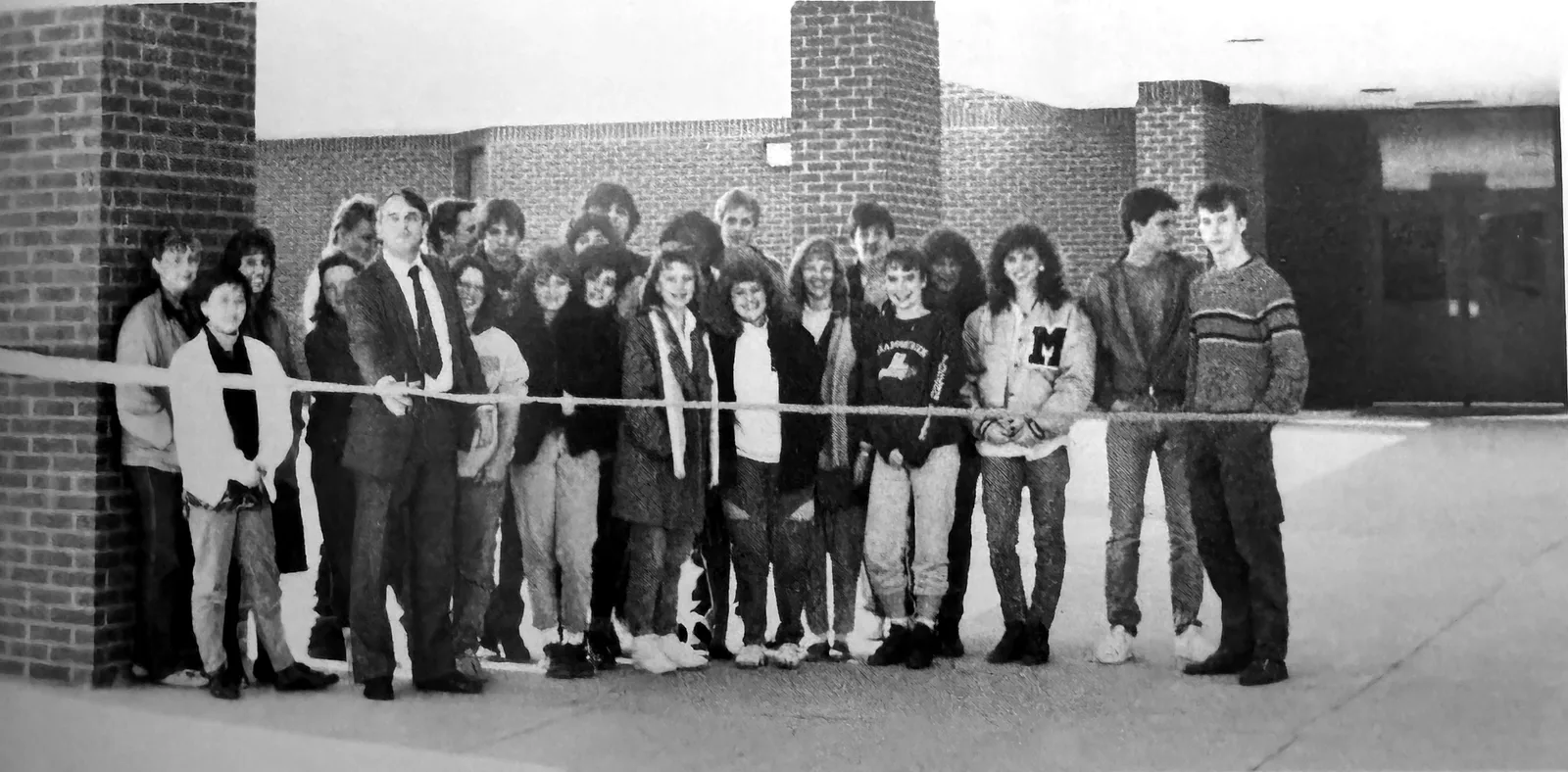Black and white yearbook photo of students, teachers, and community members lined up behind a ribbon in front of Meadowcreek High School at the close of the inaugural year
